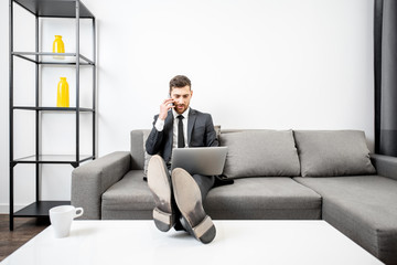 Elegant businessman dressed in the suit working with laptop and talking phone on the couch at home or comfortable office