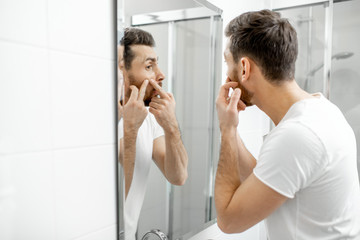 Man in white t-shirt squeezing acne on his face looking to the mirror in the bathroom