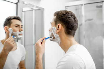 Handsome man in white t-shirt shawing his beard with blade and foam in the bathroom