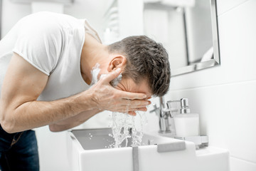 Man washing his face with fresh water and foam in the sink at the white bathroom