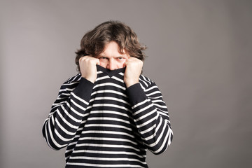 Young man pulling black and white striped sweater over head against gray background. Angry male picks up a sweatshirt over his head. Loneliness of depressed person.