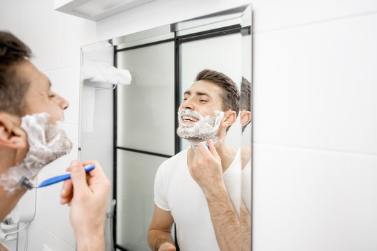 Handsome man in white t-shirt shawing his beard with blade and foam in the bathroom