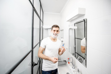 Naklejka na ściany i meble Portrait of a handsome man in white t-shirt shawing with blade and foam in the bathroom