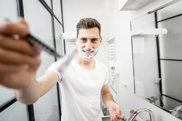 Portrait of a playful man in white t-shirt shawing with blade and foam in the bathroom