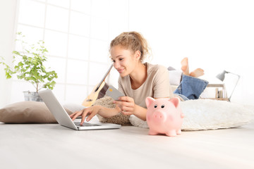 Young woman smiling holding credit card and using laptop computer. piggy bank on floor, online...