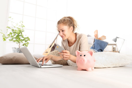 Young woman smiling holding credit card and using laptop computer. piggy bank on floor, online shopping saving concept, lying on living room wooden floor in modern home, with copy space