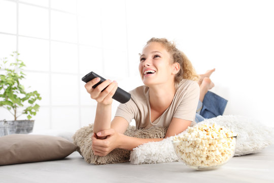 beautiful young smiling woman with tv remote control watching the television eating popcorn lying on living room wooden floor in comfortable home