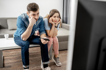 Young couple with unhappy faces watching boring TV sitting together on the table at home