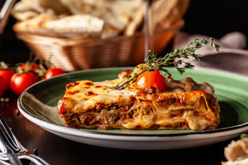 The concept of Italian cuisine. Baked lasagna with minced bolognese, pasta, cherry tomatoes lies on a green plate in a restaurant. A glass of red wine and bread basket on the table