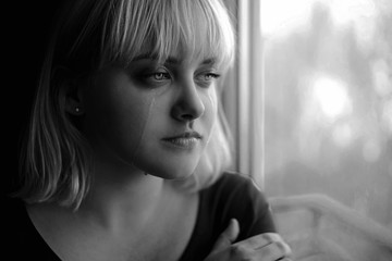 Close up of crying depressed young blond woman near window at home. Sadness, nostlagic, depression. Black and white photo.