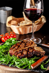 The concept of Italian cuisine. Juicy T-bone steak of beef aging stewed meat, with rucola salad and cherry tomatoes. A glass of red wine and bread basket on the table