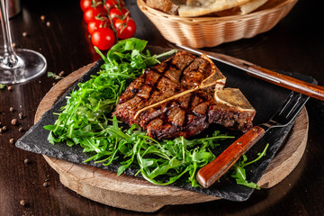 The concept of Italian cuisine. Juicy T-bone steak of beef aging stewed meat, with rucola salad and cherry tomatoes. A glass of red wine and bread basket on the table
