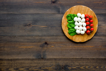 Italian flag made of mozzarella cheese, cherry tomatoes, green basil on wooden cutting board on dark wooden background top view copy space