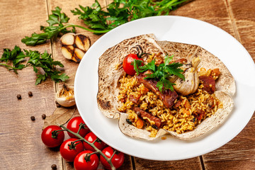 The concept of Eastern, Turkish, Uzbek cuisine. Pilaf with pieces of meat is served in pita bread, in a white plate on a wooden table. parsley and vegetables on the table.