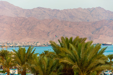 Fototapeta na wymiar View from Eilat city through palm tops on Red Sea bay and Aqaba city of Jordan with high sand mountains