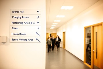 Secondary School corridor with signage to sports hall, changing rooms, performing arts, toilets, fitness room and sports viewing area with out of focus school children.