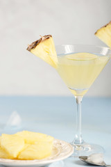 A glass of pineapple drink. The glass is decorated with slices of pineapple and lime. Next to it is a plate of fresh pineapple slices. Light background. Close-up. Vertical frame orientation.