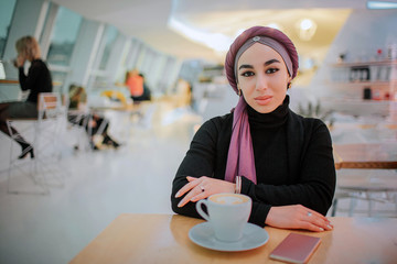 Arabian woman sits inside in cafe at table and looks on camera. She is attractive and beautiful. Cup of coffee is on table.