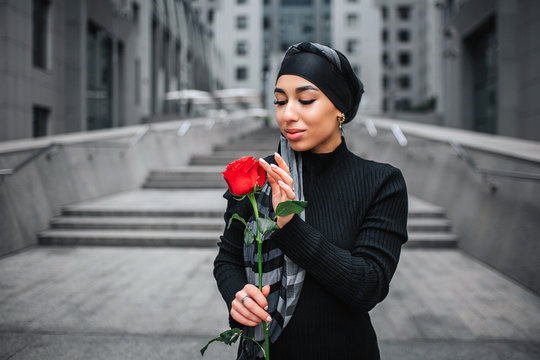 Beautiful young arabian woman hold red rose. She looks and touches it. Model smiles a bit. She stands on steps.