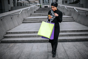 Young arabian woman walks and look at phone. She holds shopping bags. Model touches edge of her...
