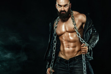 Men fashion. Close-up portrait of a brutal bearded man topless in a leather jacket with chains....