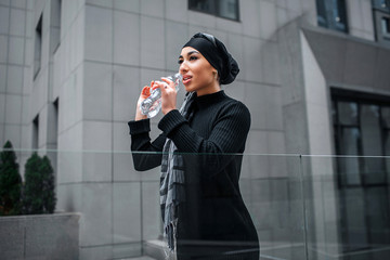 Fototapeta premium Attractive arabian woman drinks water from bottle. She looks straight forward. Woman stands at edge of glass railings.
