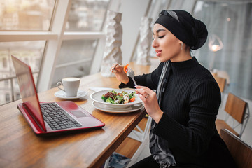 Nice young arabian woman sit at table in front of window. She eats salad. Laptop in on table.