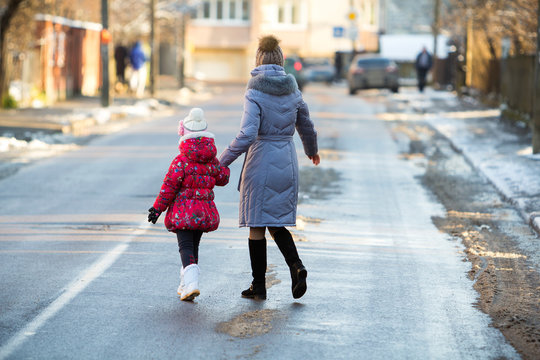 Back View Of Young Slim Attractive Woman Mother And Small Child Girl Daughter In Warm Clothing Walking Together Holding Hands Crossing Slippery Street On Sunny Winter Day On Blurred Urban Background.