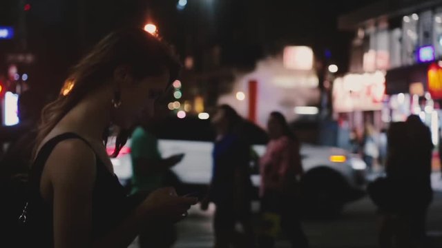 Atmospheric shot of happy Caucasian young local woman using smartphone app in dark busy New York night street crossing.