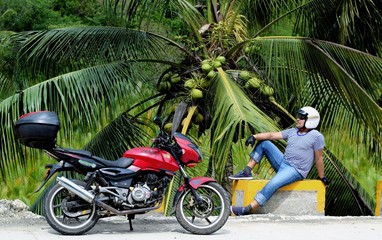 Biker with a motorcycle resting under a palm tree with coconats