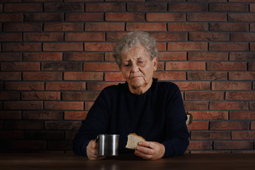 Poor elderly woman with piece of bread and metal mug at table