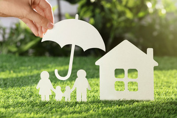 Woman holding paper umbrella over cutout of family and house on fresh grass, closeup. Life...