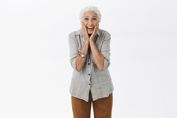 Senior woman surprised and happy like child receiving gift from children. Portrait of delighted and amused old woman with white hair touching cheeks with palms gazing amazed at camera over grey wall