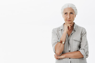 Waist-up shot fo concerned woman thinking about kids. Portrait of thoughtful upset charming senior lady with gray hair holding hand on chin looking at upper left corner, thinking over white background