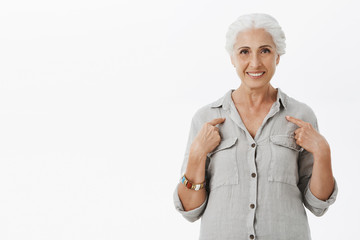 Studio shot of kind and cute elderly mother with gray hair pointing at herself with pleased and self-assured smile suggesting her candidatute feeling confident she can handle any job over white wall