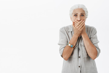 Waist-up shot of shocked and stunned elderly woman in casual shirt with grey hair gasping covering...