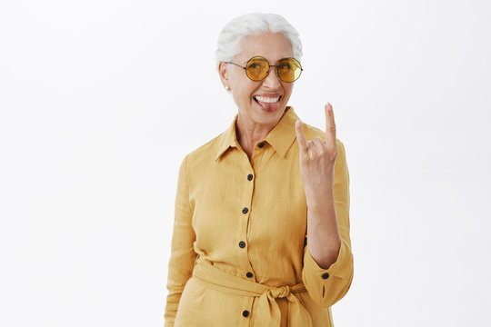 Her sould is young. Portrait of joyful pleased and carefree enthusiastic grandmother with grey hair in stylish sunglasses and yellow coat sticking out tongue showing rock n roll gesture