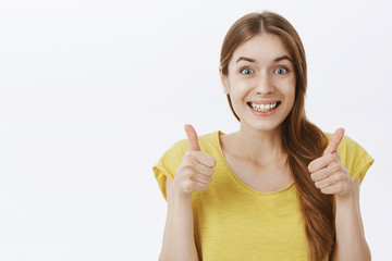 Portrait of charming excited and supportive friendly young girl with braid in yellow casual t-shirt showing thumbs up and smiling with agreement, liking idea approving good plan over gray background