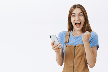Waist-up shot of triumphing charming lucky girlfriend in brown overalls raising clenched fist in joy and victory celebrating new score in game holding smartphone hazing happy at camera over grey wall