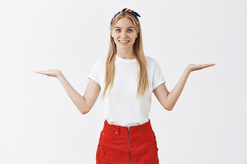 Girl gives up choice. Good-looking friendly blonde female in trendy outfit raising palms aside in weighing pose and smiling broadly at camera while making decision or offering products to compare