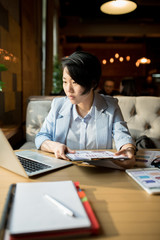 Serious concentrated attractive young Asian financial adviser in blue stripped jacket sitting at table and analyzing chart in paper while viewing information on laptop, she working in cafe