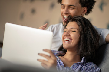 Cheerful happy African American young marriage laughing while watching movie on laptop computer at home interior.
