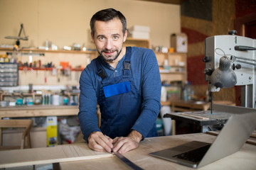 Waist up portrait of smiling mature carpenter looking at camera while preparing wood working in...