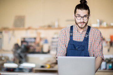 Waist up portrait of young worker using laptop and looking at camera  standing at table in modern workshop, copy space