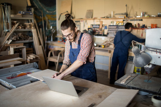 Portrait of two workers in carpenters workshop, focus on modern young man using laptop in foreground, copy space