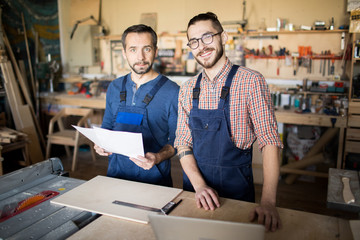 Waist up portrait of two smiling carpenters looking at camera while posing in joinery workshop,...