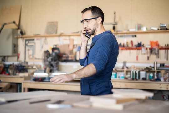 Side view portrait of mature craftsman speaking by phone while working in woodworking shop, copy space