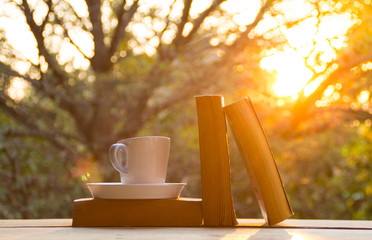 A cup of coffee, pencil and book on the wooden table with morning light and blurred nature background