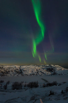 Beautiful northern lights, aurora in the night sky above the snow-covered hills. Large stones and a frozen lake.