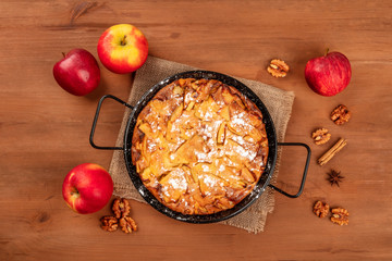 An apple pie in a pan, shot from the top on a dark rustic wooden background with apples, walnuts, cinnamon, anise, and copy space